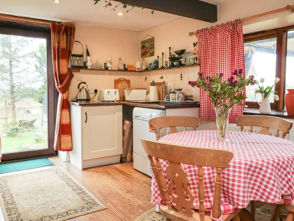 A cozy kitchen with a dining table set for four, a window with curtains, and various kitchen appliances. - Barley Meadow (Okehampton)