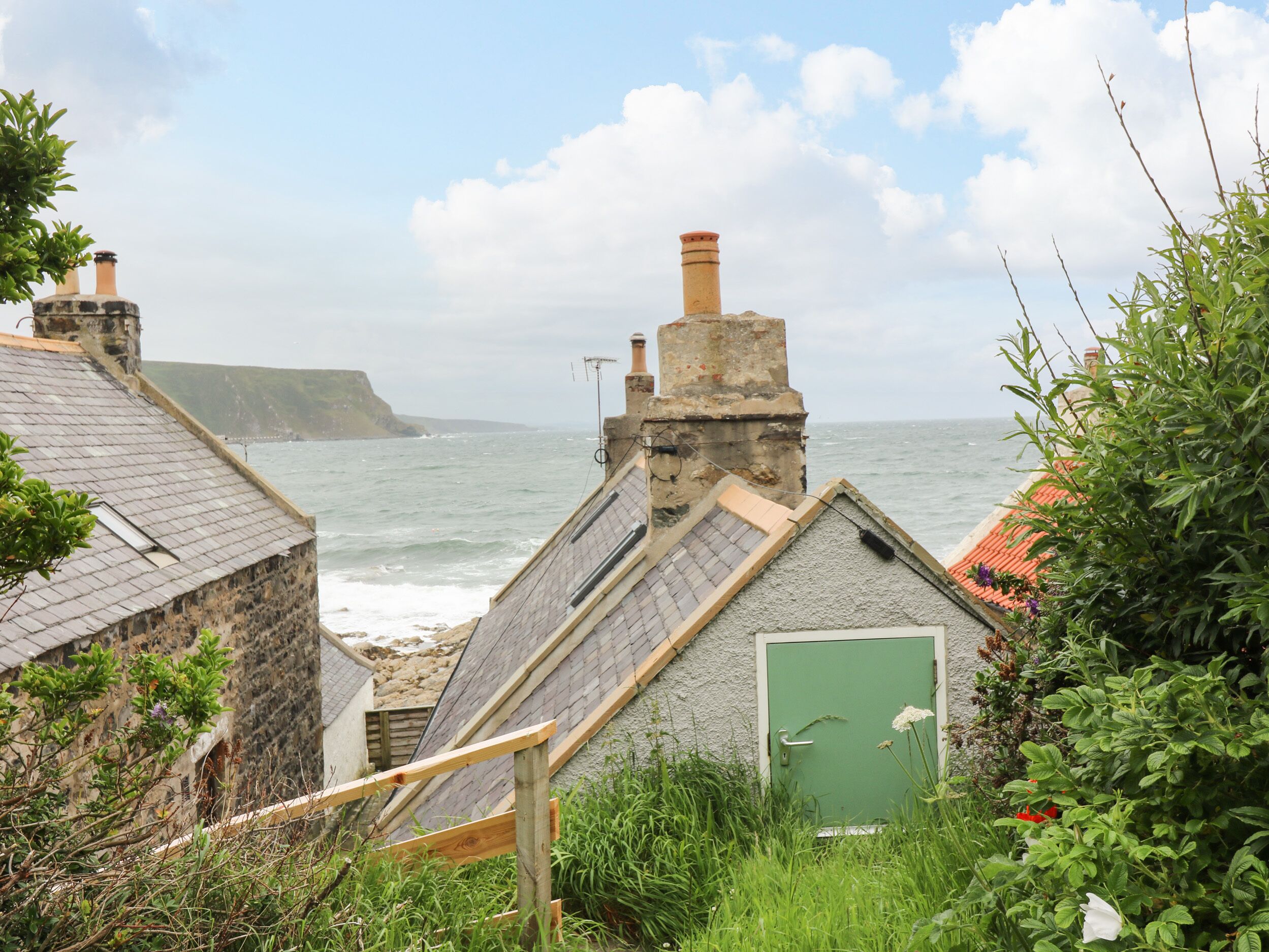 Um edifício costeiro com telhado inclinado, porta verde e vista para o mar e falésias.