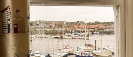 Vista de uma marina com barcos atracados, uma cidade ao fundo e um prédio com porta vermelha e um sino.