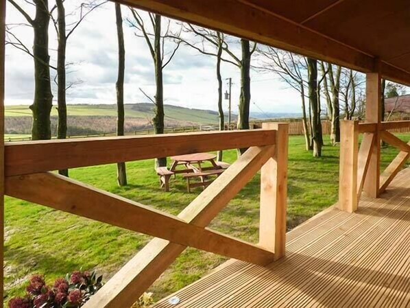 A wooden deck with a picnic table and benches, overlooking a grassy area with trees and hills. - Valley View Lodge (Welshpool)
