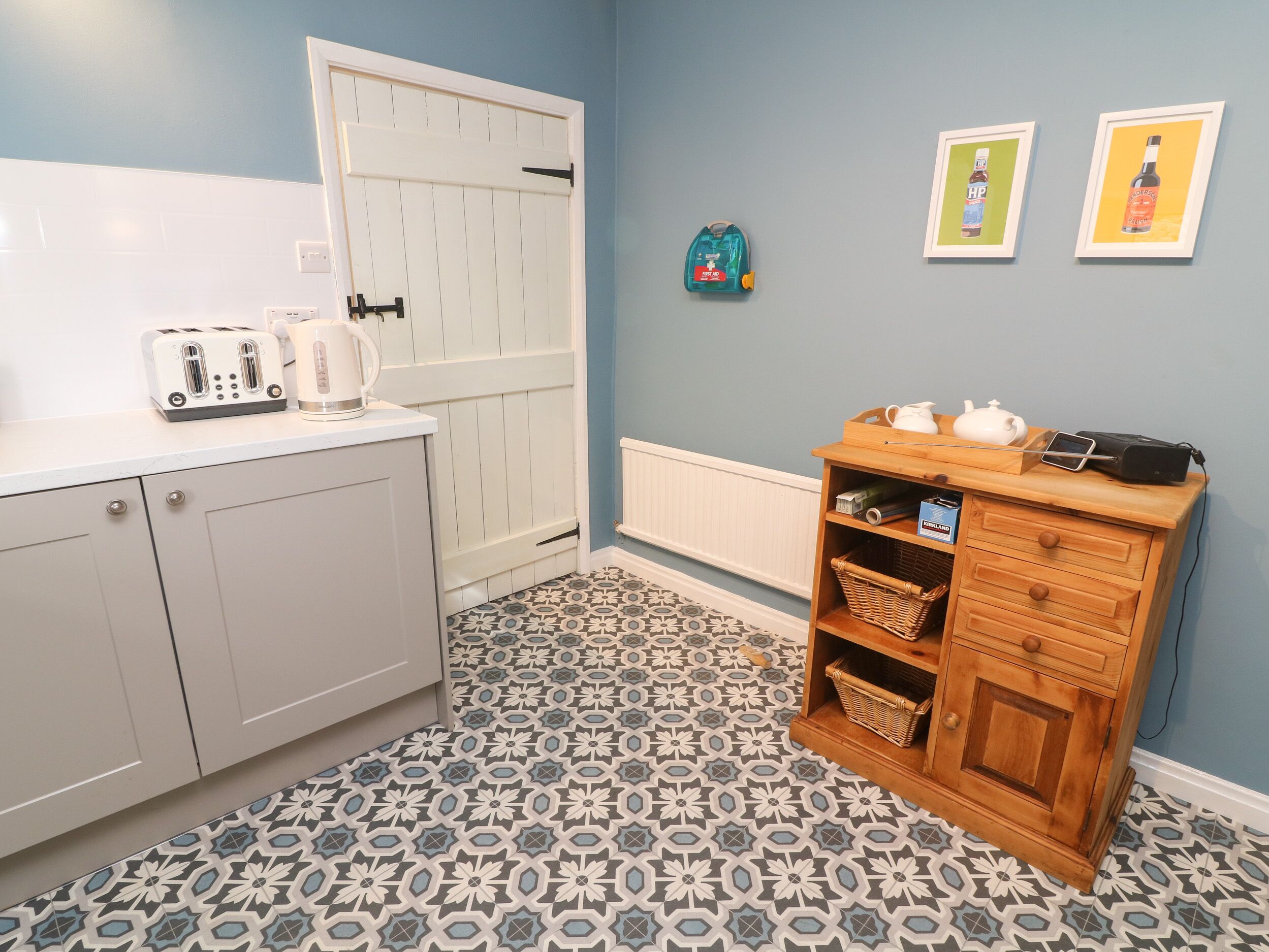 Cocina compacta con armarios blancos, piso estampado, mueble de madera con cestas y cuadros en la pared.
