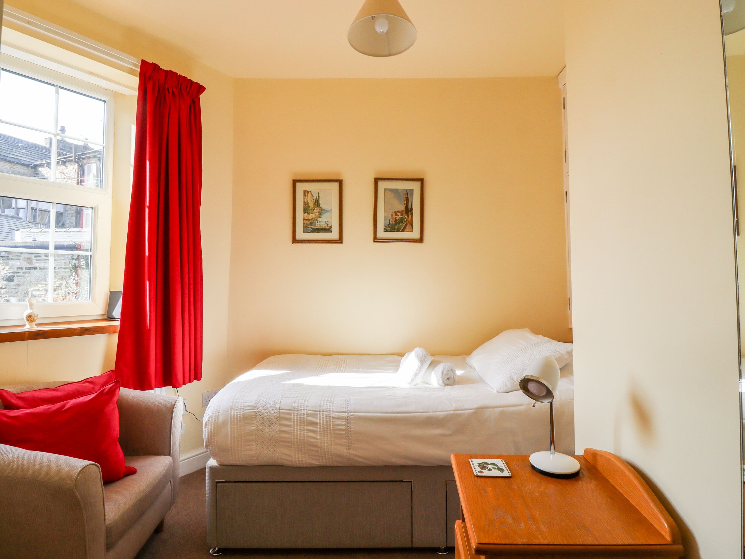 A hotel room with a bed, a bedside table with a lamp, a window with a red curtain, and two framed pictures on the wall.