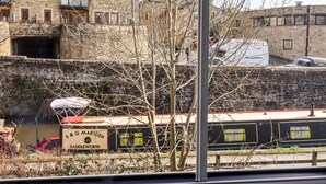 Vista de un edificio de piedra con varias ventanas y balcones, un estrecho canal con una barca, y un árbol en primer plano.