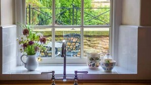 Ein Küchenfenster mit Blick auf einen Garten, ein weißer Krug mit Blumen, ein Wasserhahn und ein Küchengeschirr-Set.