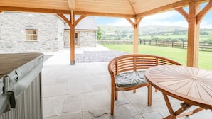 Un patio techado con suelo de piedra, una mesa y silla de madera, y un edificio de piedra al fondo.