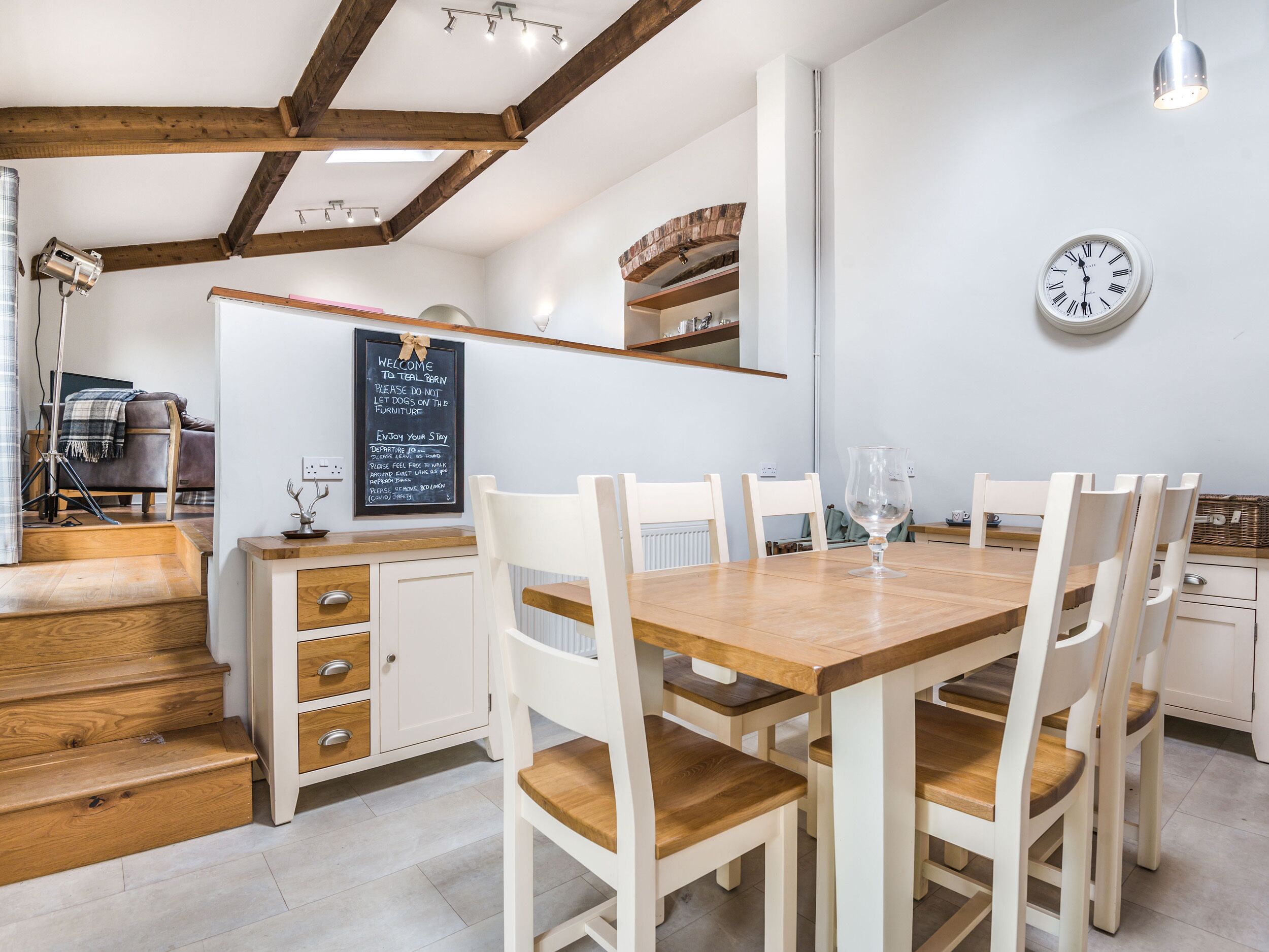 Un comedor con mesa de madera y sillas blancas, un reloj en la pared y una pizarra con un mensaje de bienvenida.