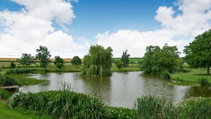 A serene pond surrounded by lush greenery and trees, with a wooden bench and a grassy area in the foreground. - Teal Barn Annexe (Hereford)