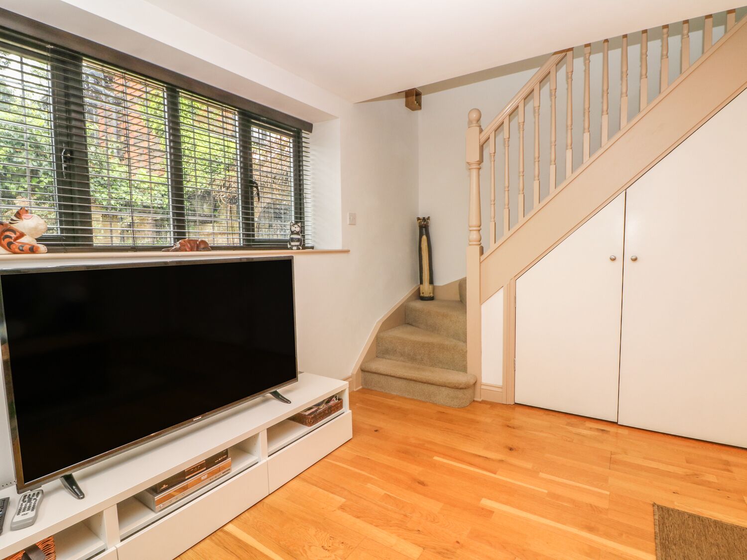 A living room with a flat-screen TV on a white entertainment unit, a staircase with a wooden handrail, and a window with blinds.