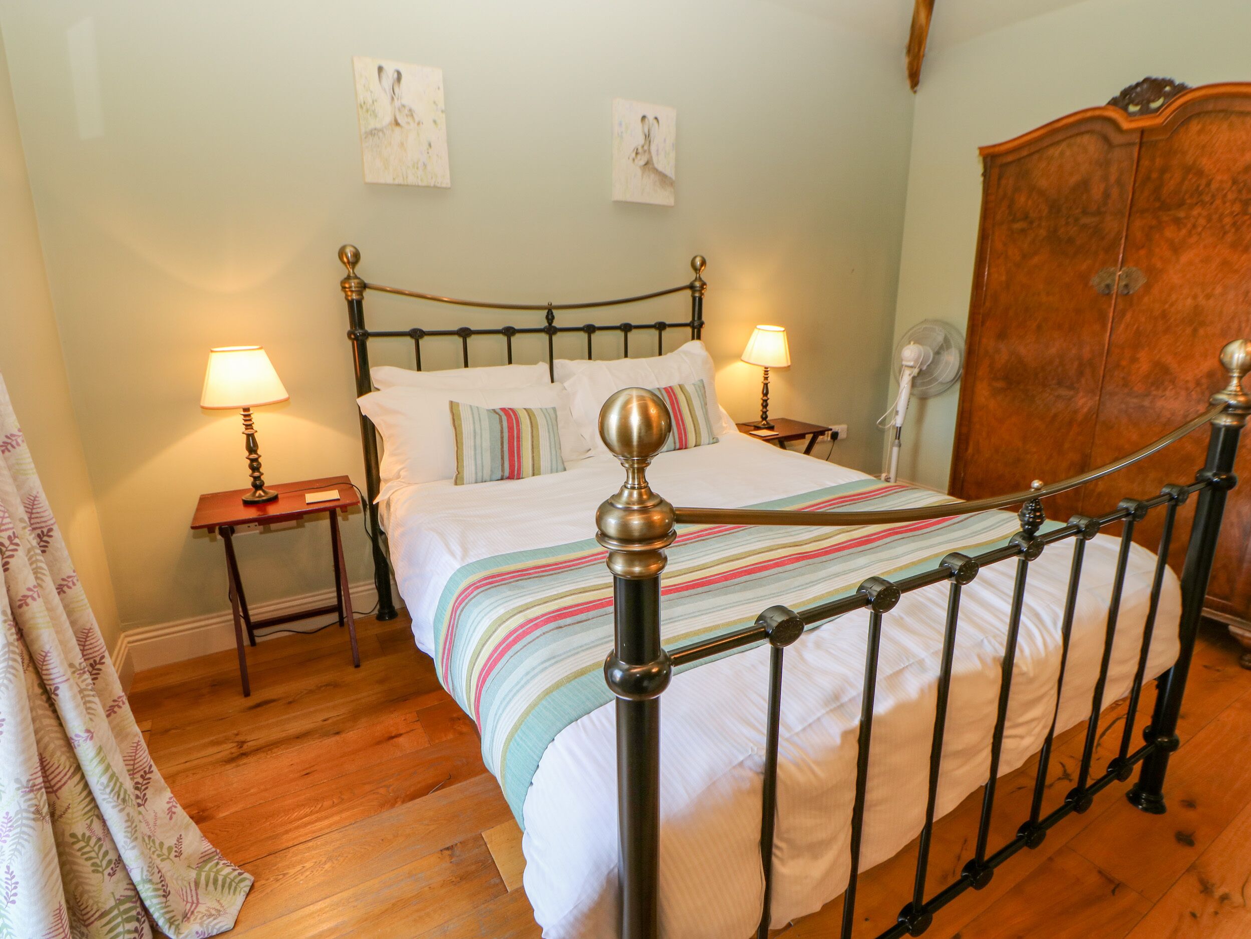 A neatly made bed with a striped blanket, two bedside lamps, and a wooden wardrobe in a room with wooden flooring.
