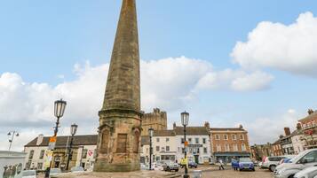 A stone obelisk monument in a town square with cobblestone streets, historic buildings, and parked cars.