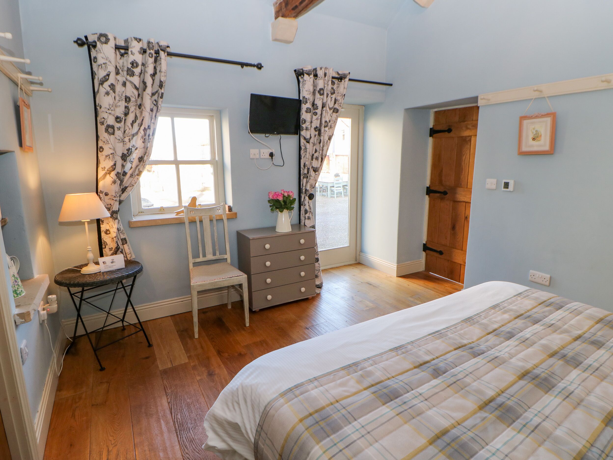 A bedroom with a bed, a dresser, a chair, a window with curtains, a television mounted on the wall, and a wooden door.
