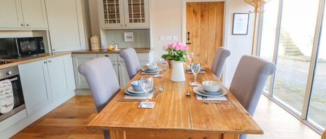 A dining area with a wooden table set for four, surrounded by light grey upholstered chairs. A white vase with pink flowers is on the table.