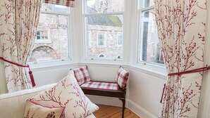 A cozy corner with a bench and cushions, large windows with curtains, and a view of buildings outside. - Iona 10 Palace Street East (Berwick-upon-Tweed)