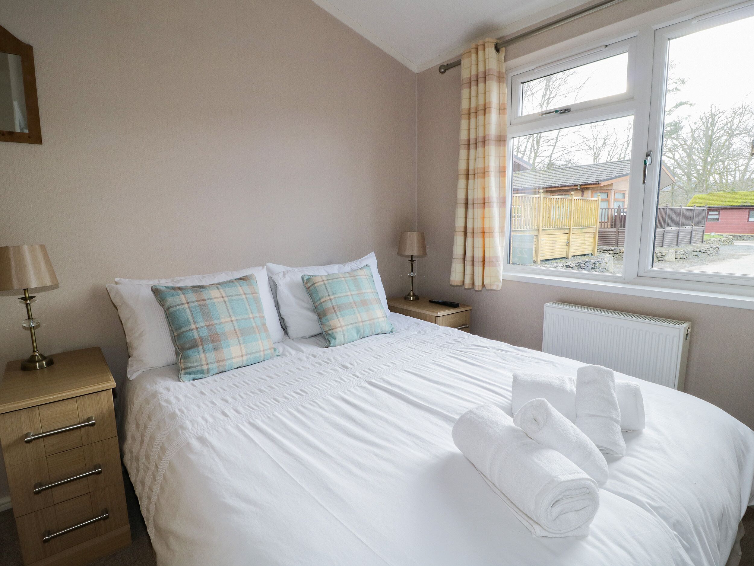 A neatly made bed with white linens and plaid pillows, a bedside table with lamps, and a window with curtains.