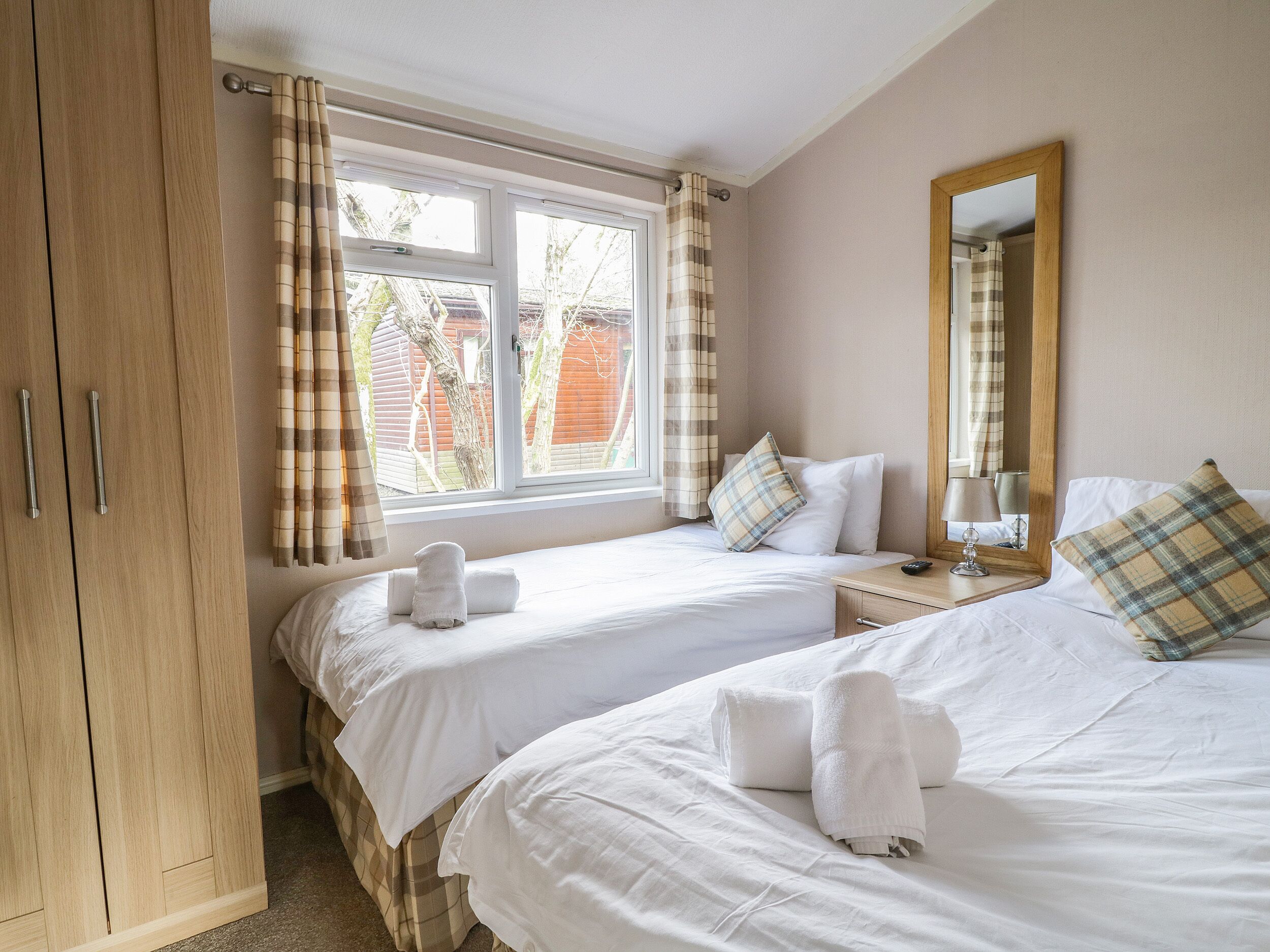 Une chambre d’hôtel avec deux lits, une armoire, une fenêtre avec des rideaux, une table de chevet avec une lampe et un miroir au mur.
