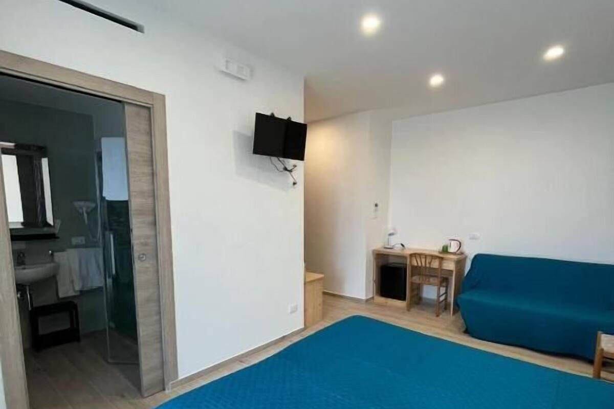 Un dormitorio con una cama grande, un escritorio con una silla, un televisor montado en la pared y un baño visible a través de una puerta abierta.