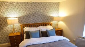 Un dormitorio con papel tapiz estampado, cabecera de madera, cama con ropa de cama blanca y azul, dos mesitas de noche con lámparas y un radiador.