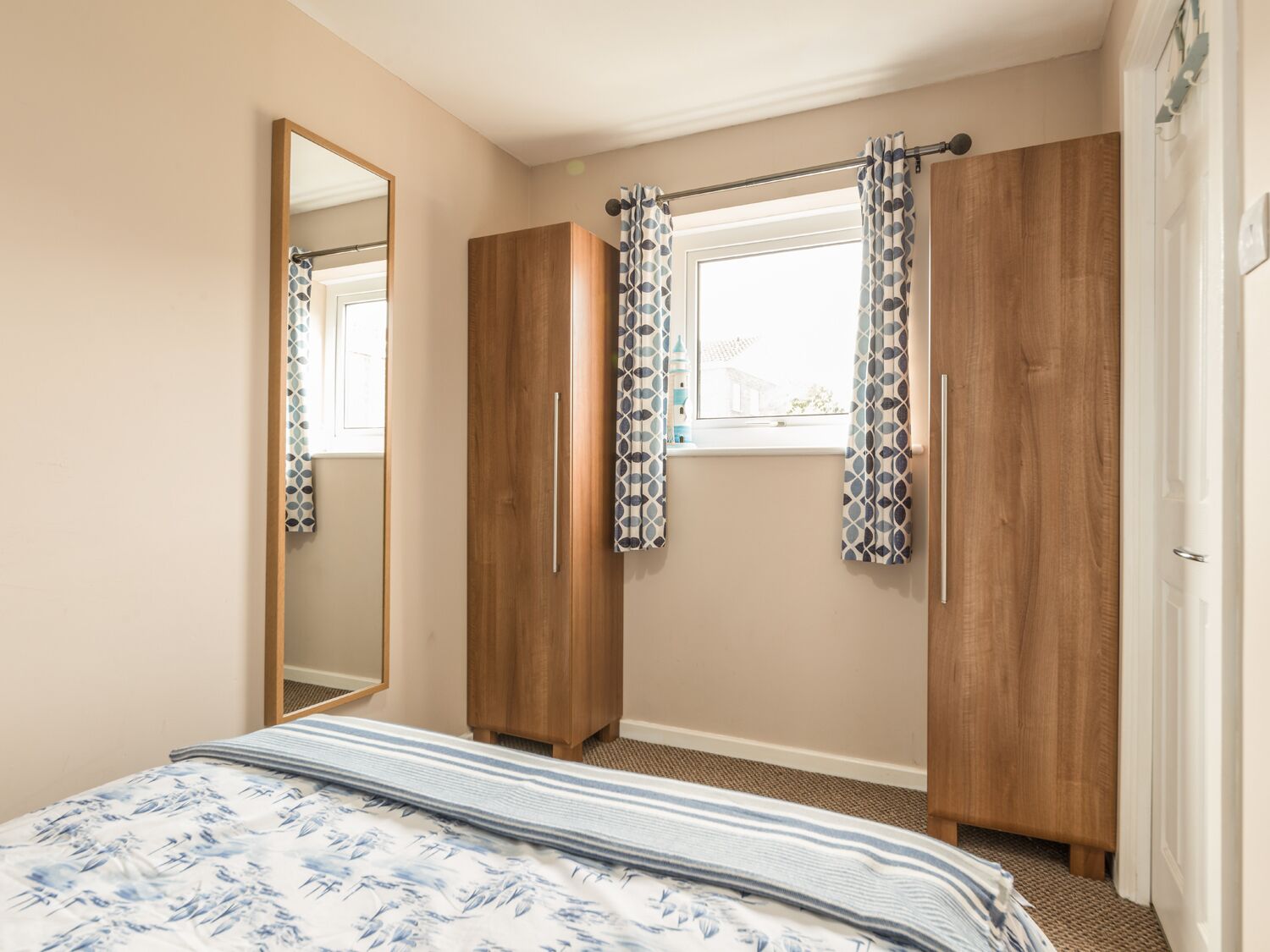 A bedroom with a bed, wooden wardrobe, a window with patterned curtains, and a mirror.