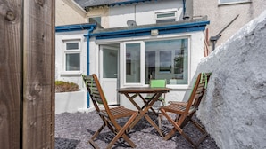 An outdoor seating area with a table and chairs, a white wall, and a window with a view of a house. - 2 Tregof Terrace (Cemaes Bay)