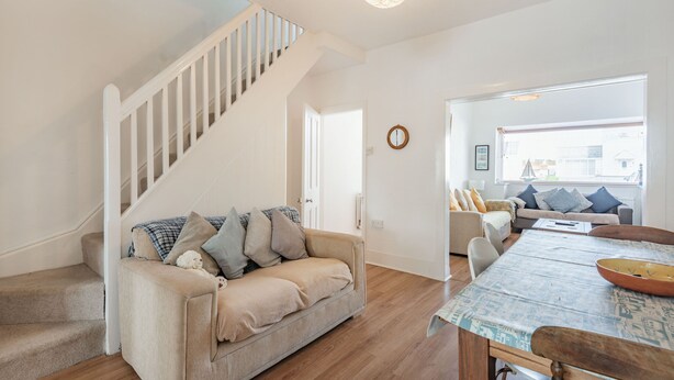 A living room with a staircase, a sofa with cushions, a dining table with chairs, and a view through a window. - 2 Tregof Terrace (Cemaes Bay)