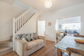A living room with a staircase, a sofa with cushions, a dining table with chairs, and a view through a window.