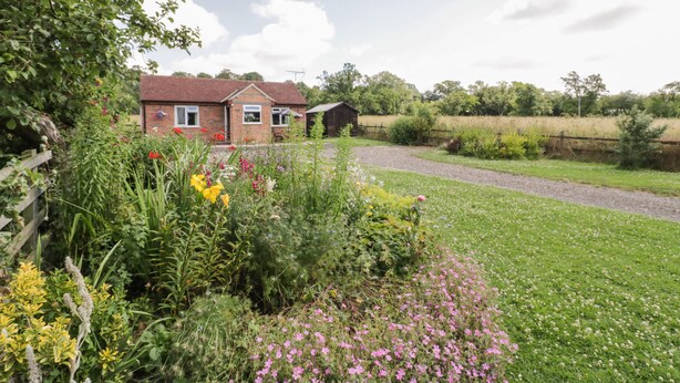 A house with a red roof, surrounded by a garden with various flowers and a gravel path leading to it. - Lawn Lodge (Gloucester)
