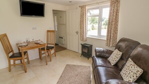 A living room with a brown leather sofa, a wooden dining table with chairs, a television mounted on the wall, and a window with patterned curtains. - Lawn Lodge (Gloucester)