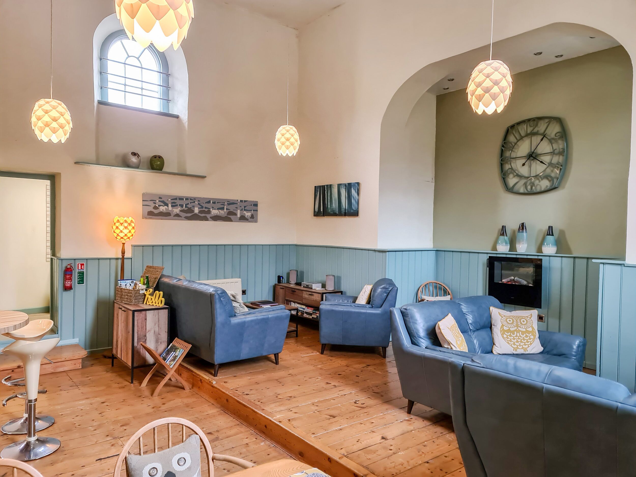 A spacious living room with blue sofas, wooden flooring, and hanging pendant lights.