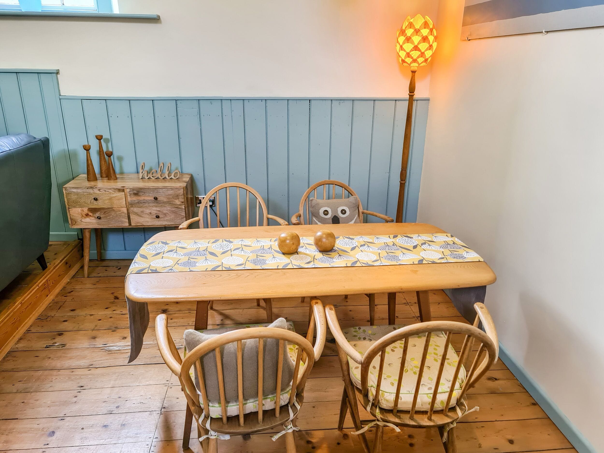 Una mesa de comedor de madera con mantel estampado, rodeada de sillas de madera con cojines. Un aparador de madera con objetos decorativos está apoyado en la pared.