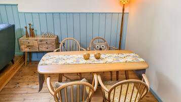 Una mesa de comedor de madera con mantel estampado, rodeada de sillas de madera con cojines. Un aparador de madera con objetos decorativos está apoyado en la pared.