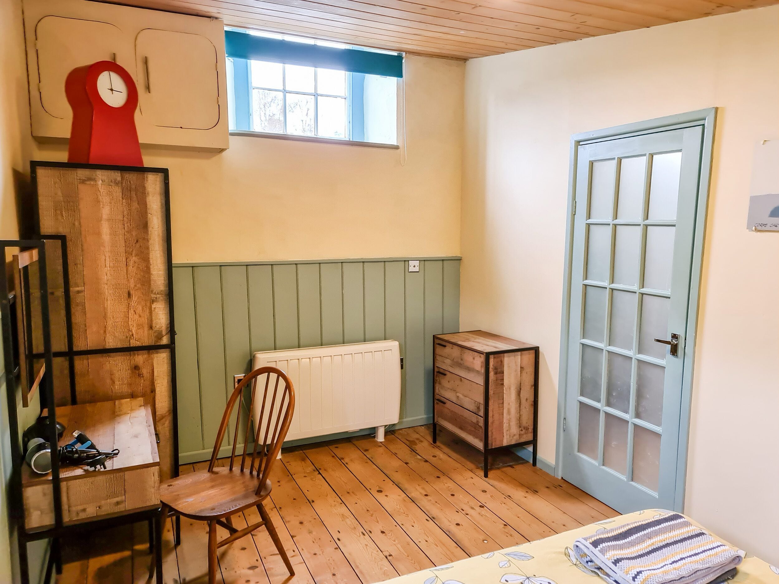 Ein Zimmer mit Holzboden, einem Heizkörper, einem Holzsessel, einem kleinen Holzkasten, einer roten Uhr und einer blauen Tür.
