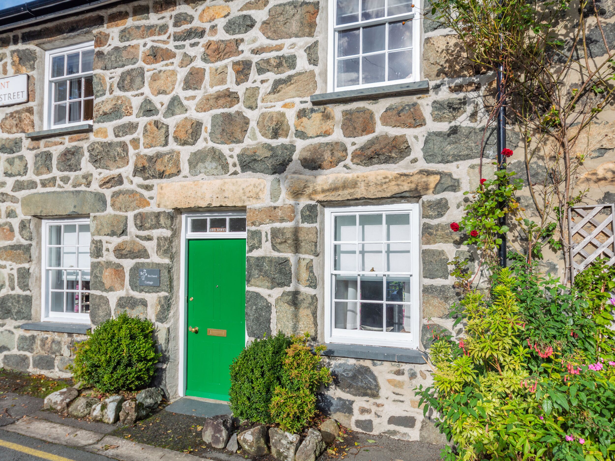 A stone building with a green door, multiple windows, and a small garden with bushes and flowers.