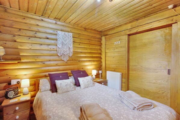 Un dormitorio de cabaña de madera con cama, mesitas de noche, una radio y un adorno macramé colgado en la pared.