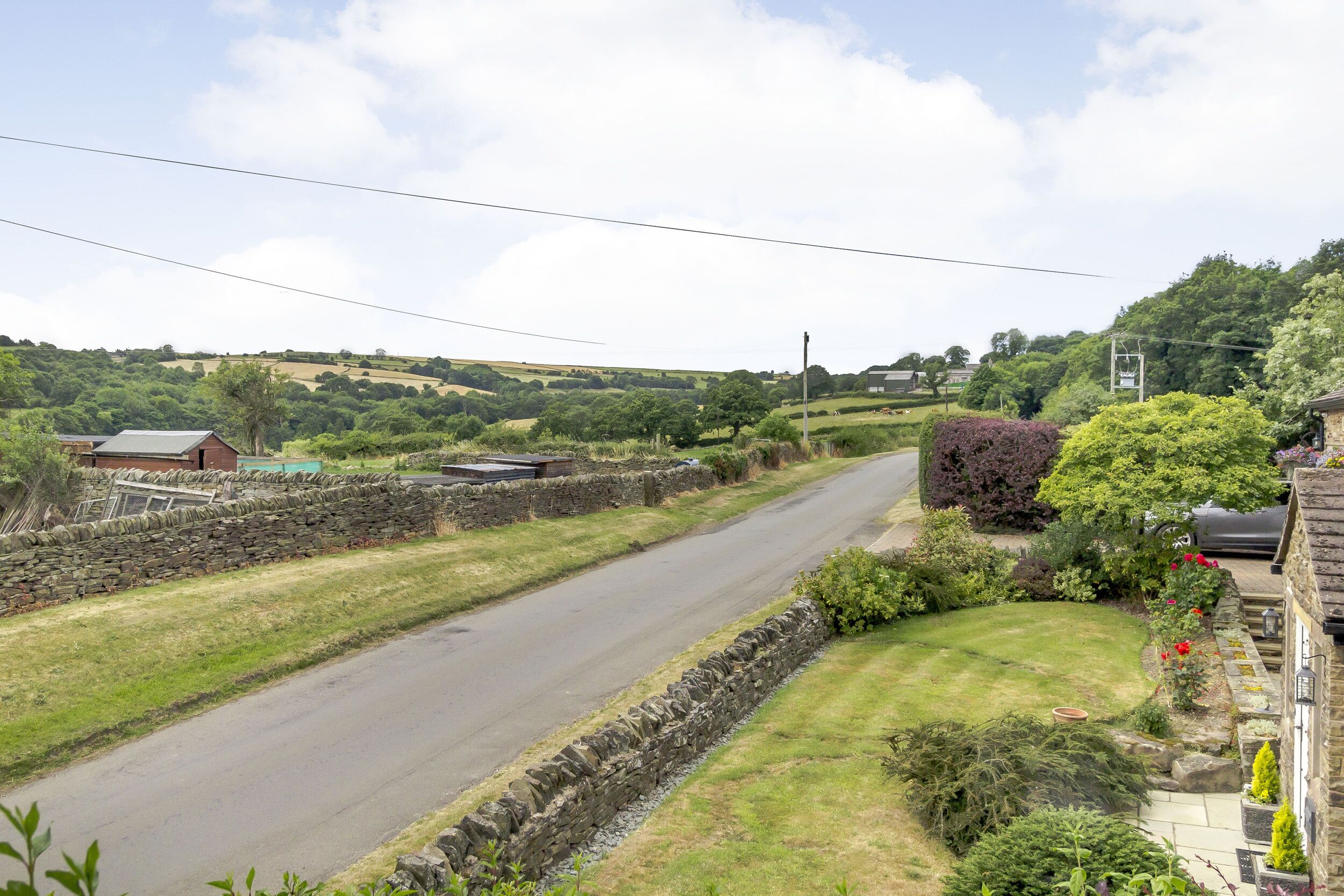 A rural road flanked by stone walls and lush greenery, with houses and a clear sky.