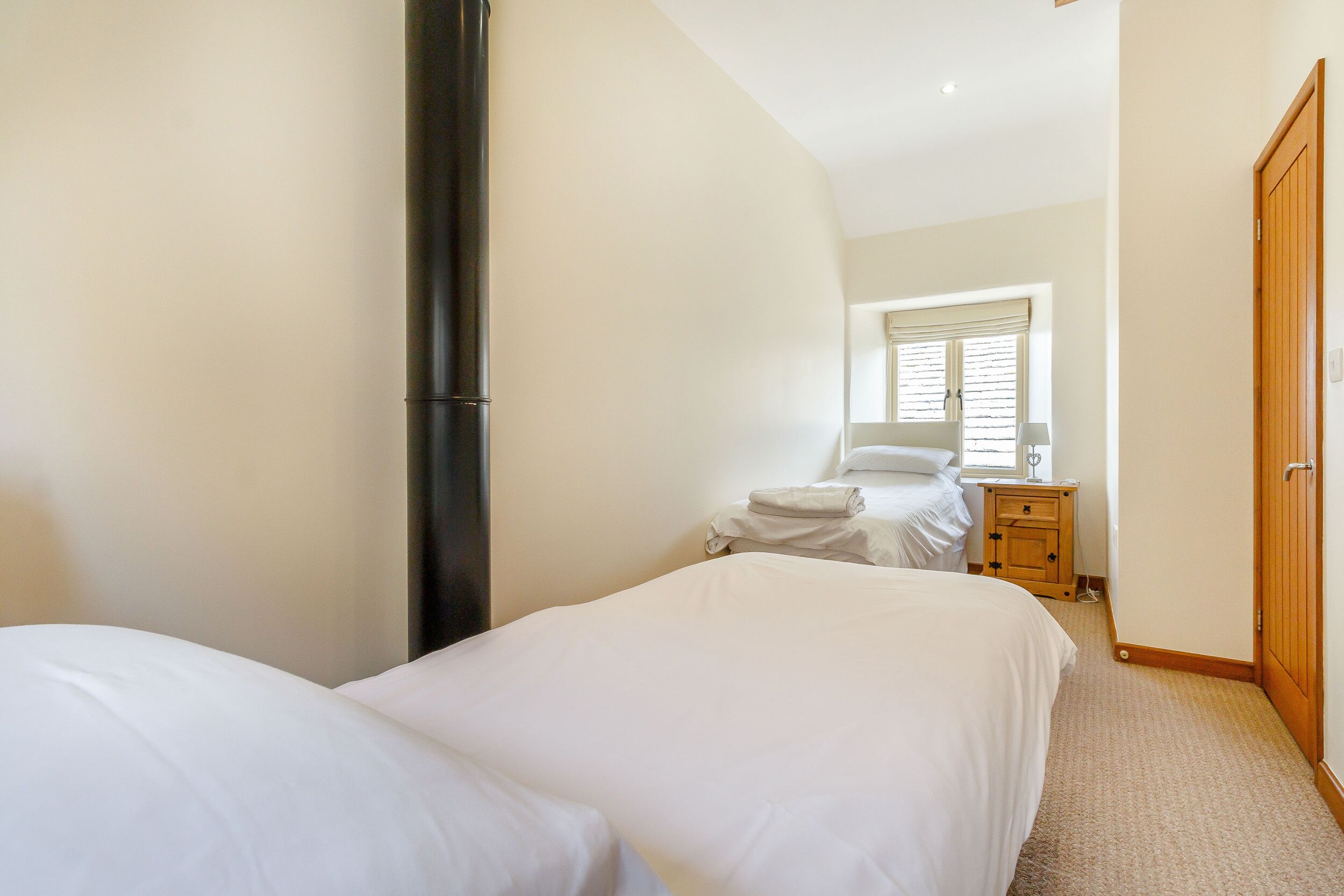 A bedroom with two beds, a wooden wardrobe, a bedside table, and a window with blinds.