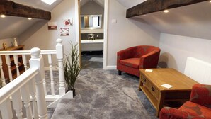 A cozy loft bedroom with a sloped ceiling, a wooden ceiling beam, a red armchair, a small wooden table, and a potted plant. - Little Owl Barn (Buxton)
