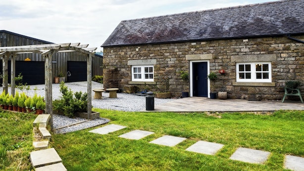 A stone building with a dark roof, a wooden pergola, and a gravel pathway leading to the entrance. - Little Owl Barn (Buxton)