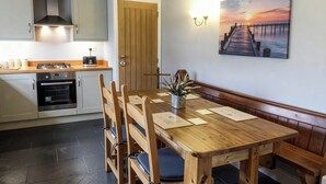 A dining area with a wooden table and benches, a kitchen with modern appliances, and a sunset painting on the wall. - Little Owl Barn (Buxton)