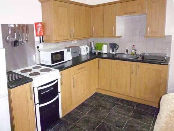 A small kitchen with wooden cabinets, a stove, a microwave, a sink, and a tiled floor. - Harley Apartment (Bishops Castle)