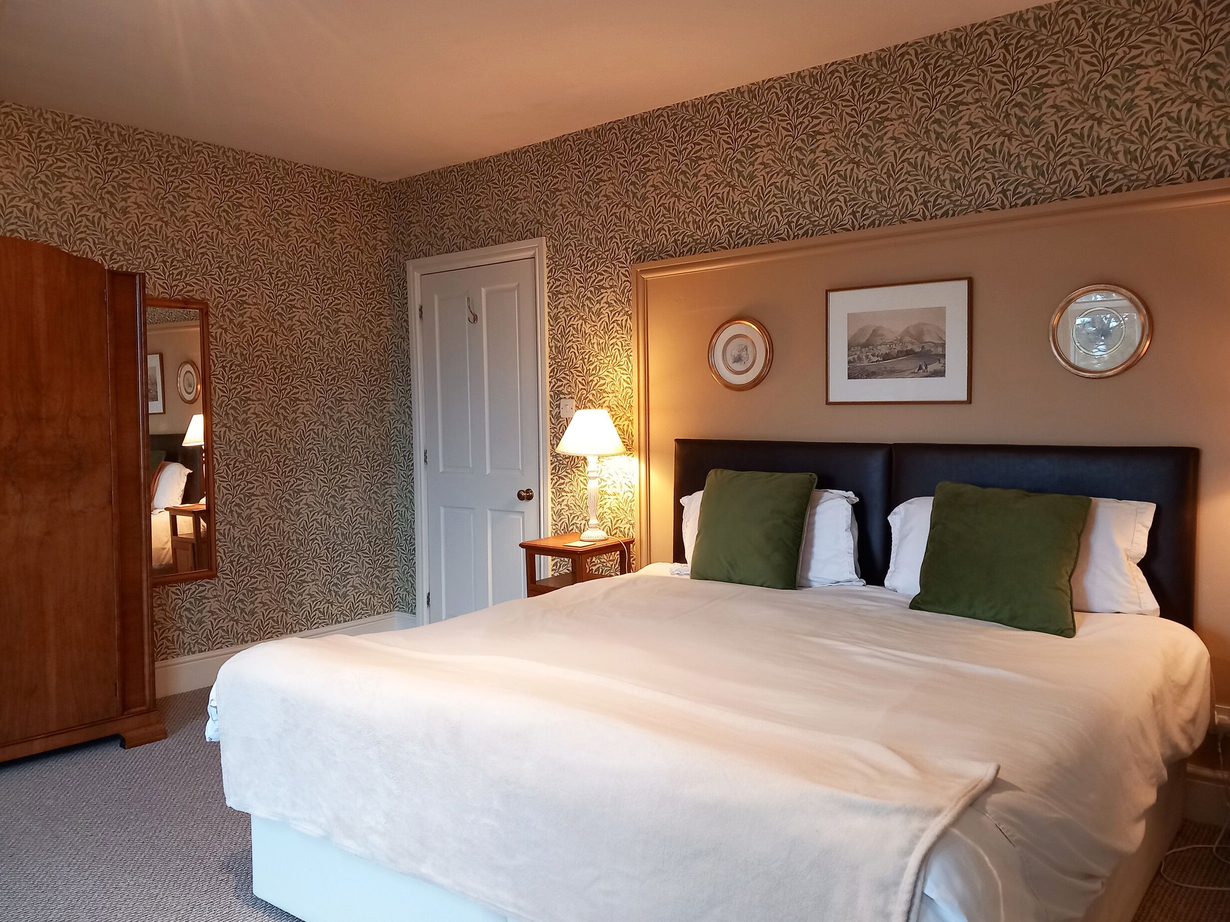 A hotel room with a large bed, a nightstand with a lamp, a wardrobe, and patterned wallpaper.