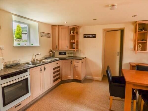 A compact kitchen with a built-in oven, a sink, and wooden cabinets. There is a dining table with chairs and a small window with a view of greenery. - Poacher's Rest (Matlock)