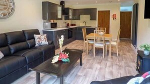 A modern living room with a black leather sofa, a coffee table with a floral centerpiece, and a dining area with wooden chairs and a table. - Cwm Gran Meadows (Pontyclun)