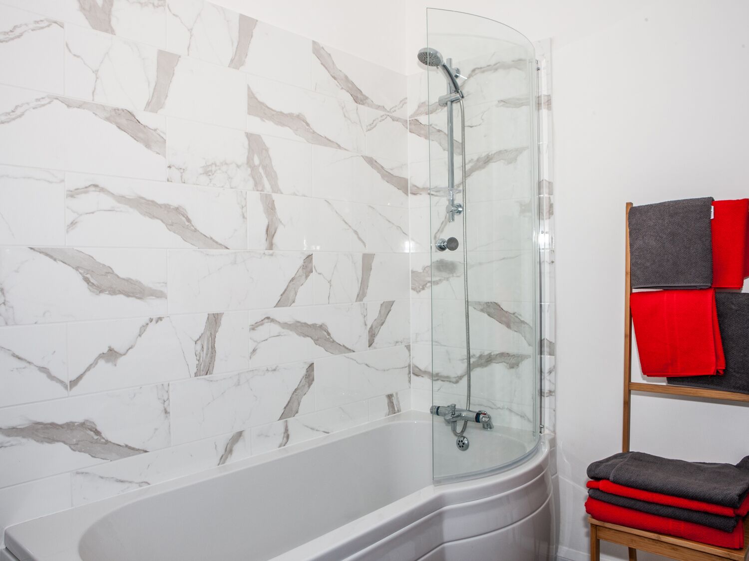 A modern bathroom with a curved glass shower enclosure, a white bathtub, and a wooden shelf with folded towels.