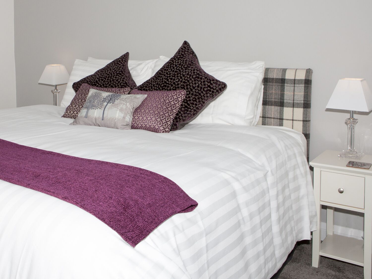 A neatly made bed with white linens and a purple throw blanket. There are two pillows with a leopard print design and one with a geometric pattern. The room has a white nightstand with a lamp and a small drawer.