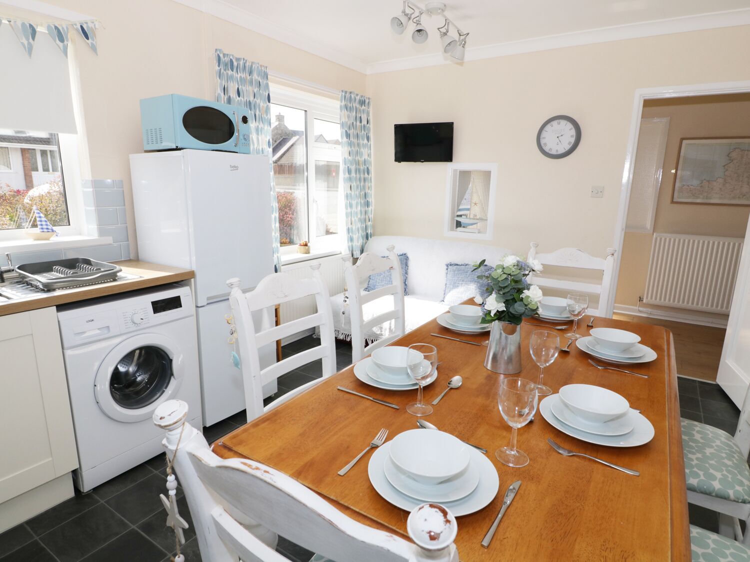 Une cuisine compacte équipée d’un lave-linge, d’un four à micro-ondes et d’un coin repas pouvant accueillir quatre personnes, avec des chaises blanches et une table en bois.