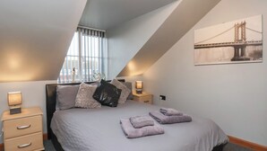 Un dormitorio con techo inclinado, una cama con almohadas y toallas, lámparas de noche, una ventana con persianas y un cuadro de un paisaje urbano en la pared.
