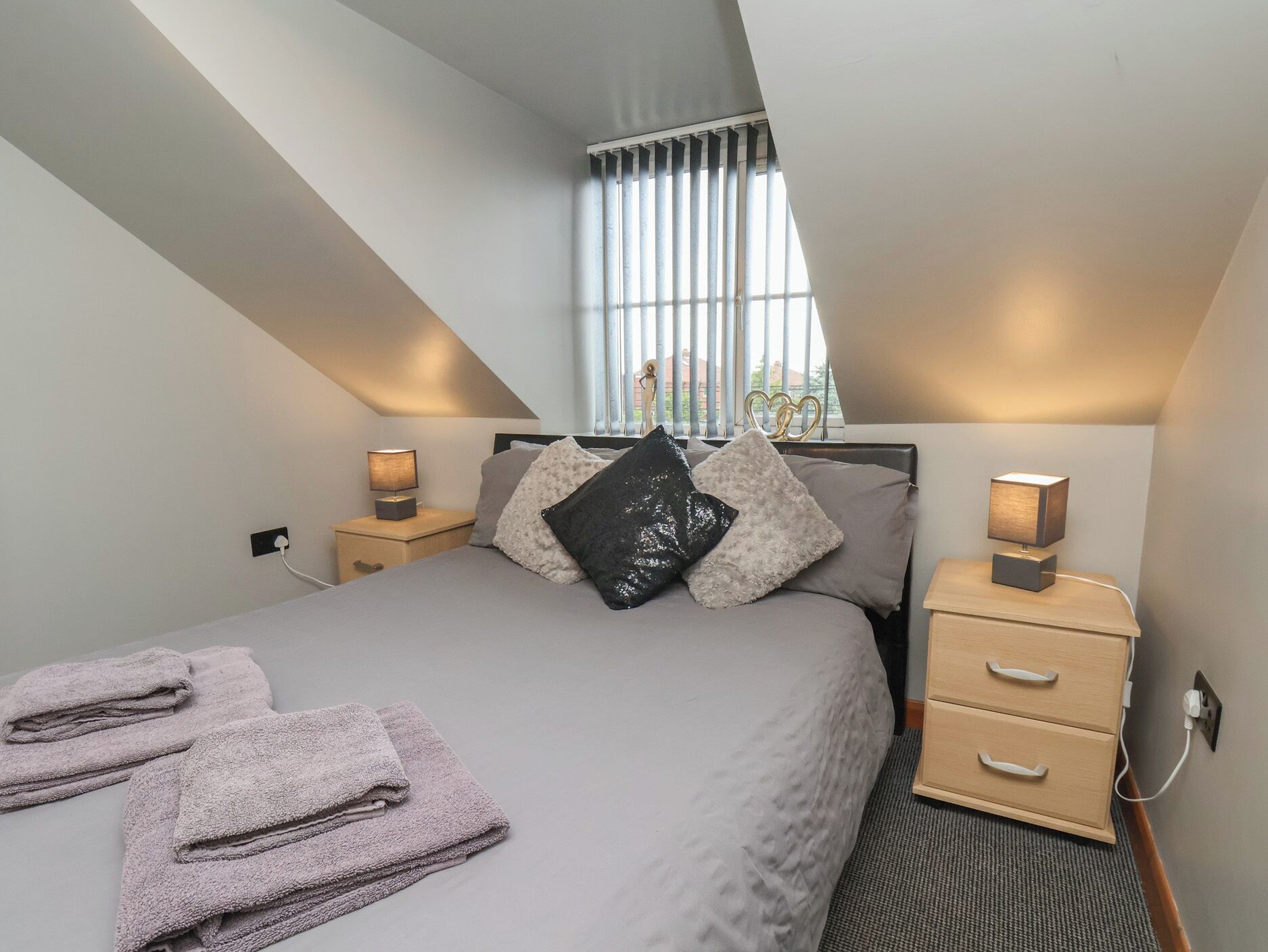 Een moderne slaapkamer met een schuine kap, een bed met veel kussens, nachtkastjes met lampen en uitzicht door het raam.