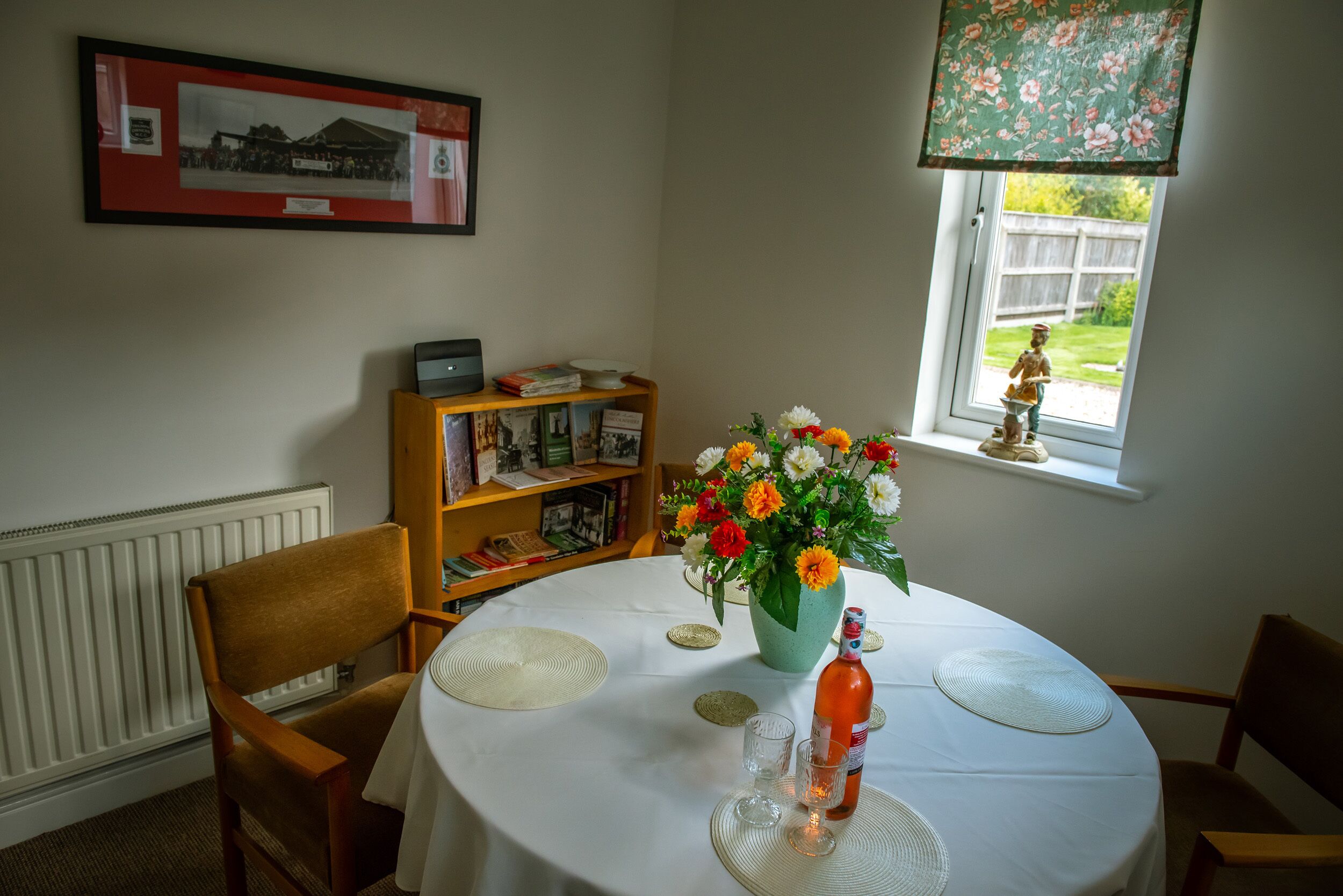 A dining table with a white tablecloth, a vase of flowers, and a bottle of wine. There is a bookshelf with books and a framed picture on the wall.