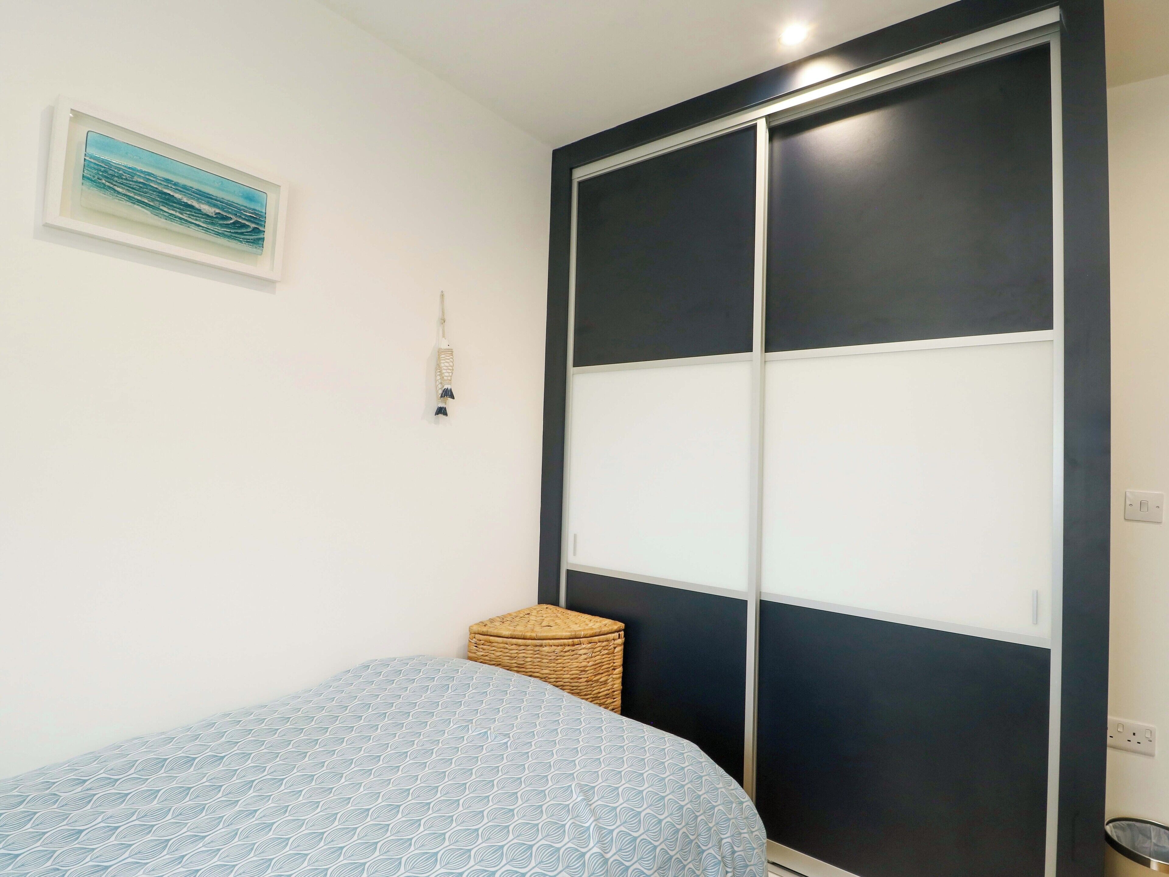 A bedroom with a bed, a wardrobe, a small woven stool, and a wall-mounted light fixture.