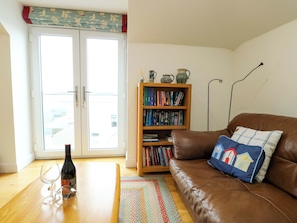A living room with a brown leather sofa, a bookshelf, a glass door with curtains, and a bottle of wine on a wooden table.